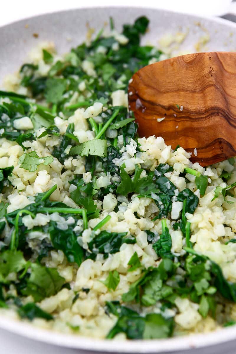 Cauliflower risotto with garlic and spinach in a white skillet being stirred with a wooden spoon.
