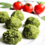 Spinach balls on a white plate with tomatoes in the background.