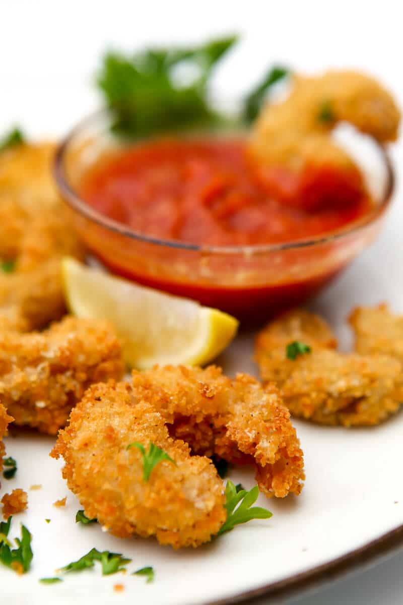 Battered and fried vegan shrimp on a white plate with a small bowl of cocktail sauce in the background.
