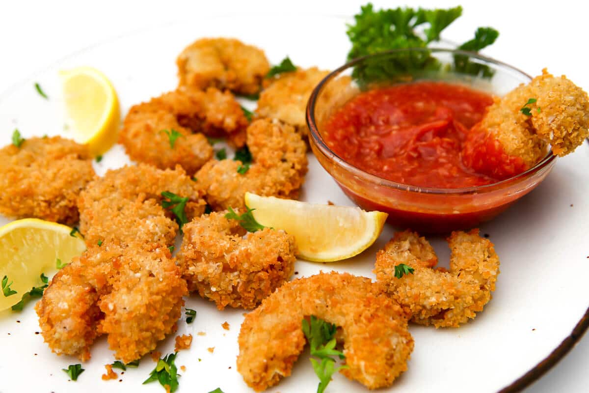A white plate full of deep fried vegan shrimp breaded with panko bread crumbs with lemon wedges and parsley.