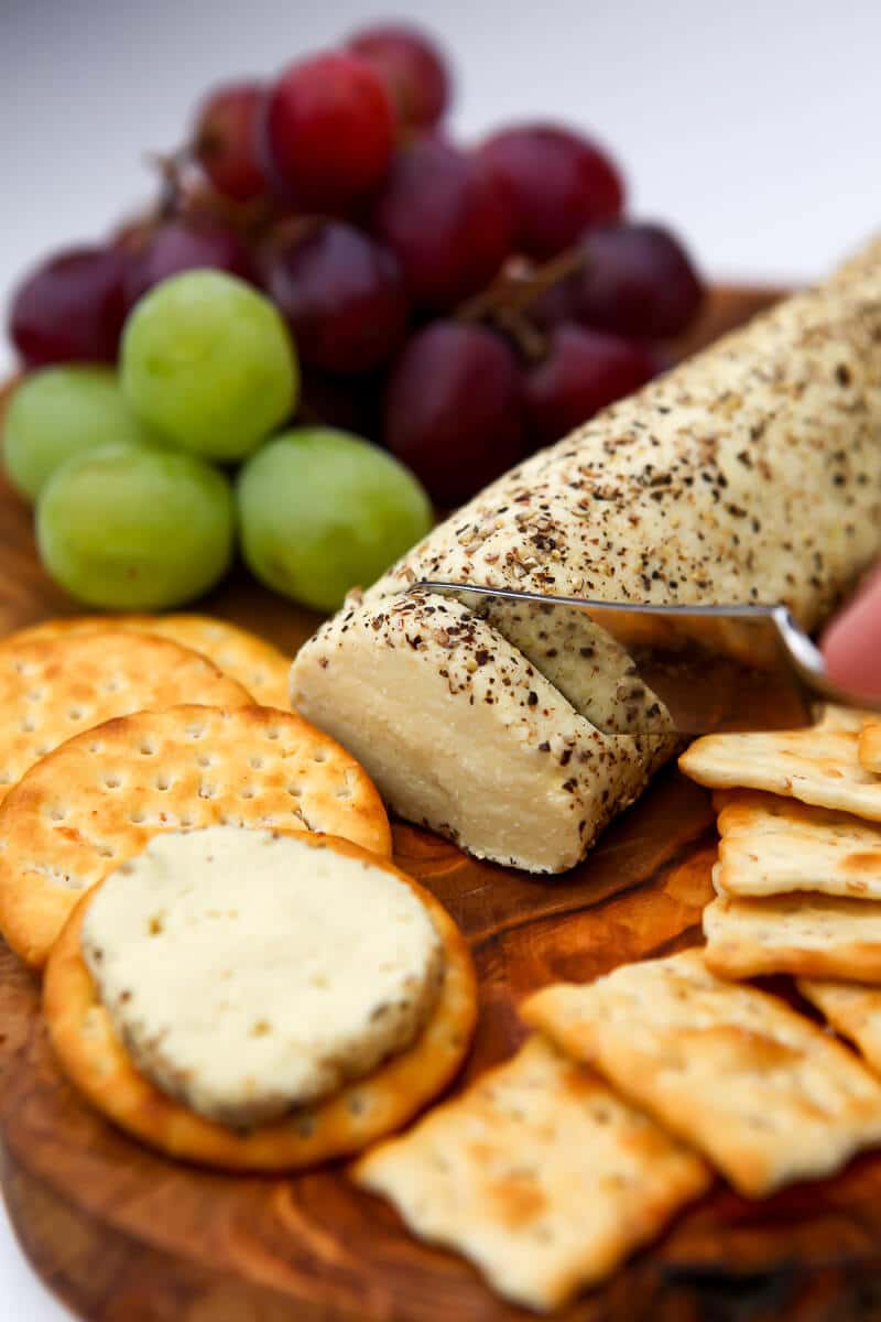 A roll of vegan goat cheese on a cutting board with grapes and crackers being cut.