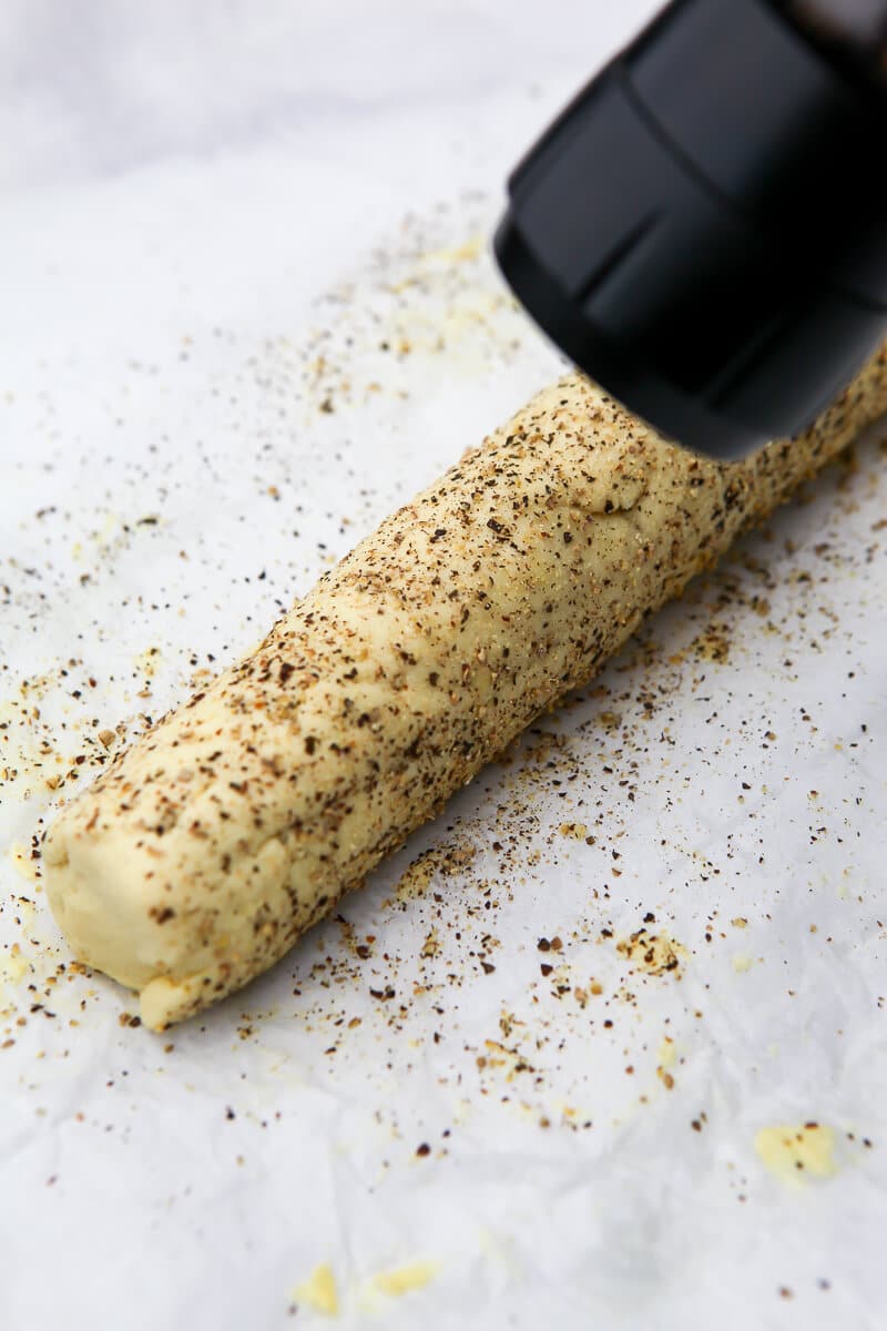 A roll of vegan goat cheese being coated in cracked pepper.