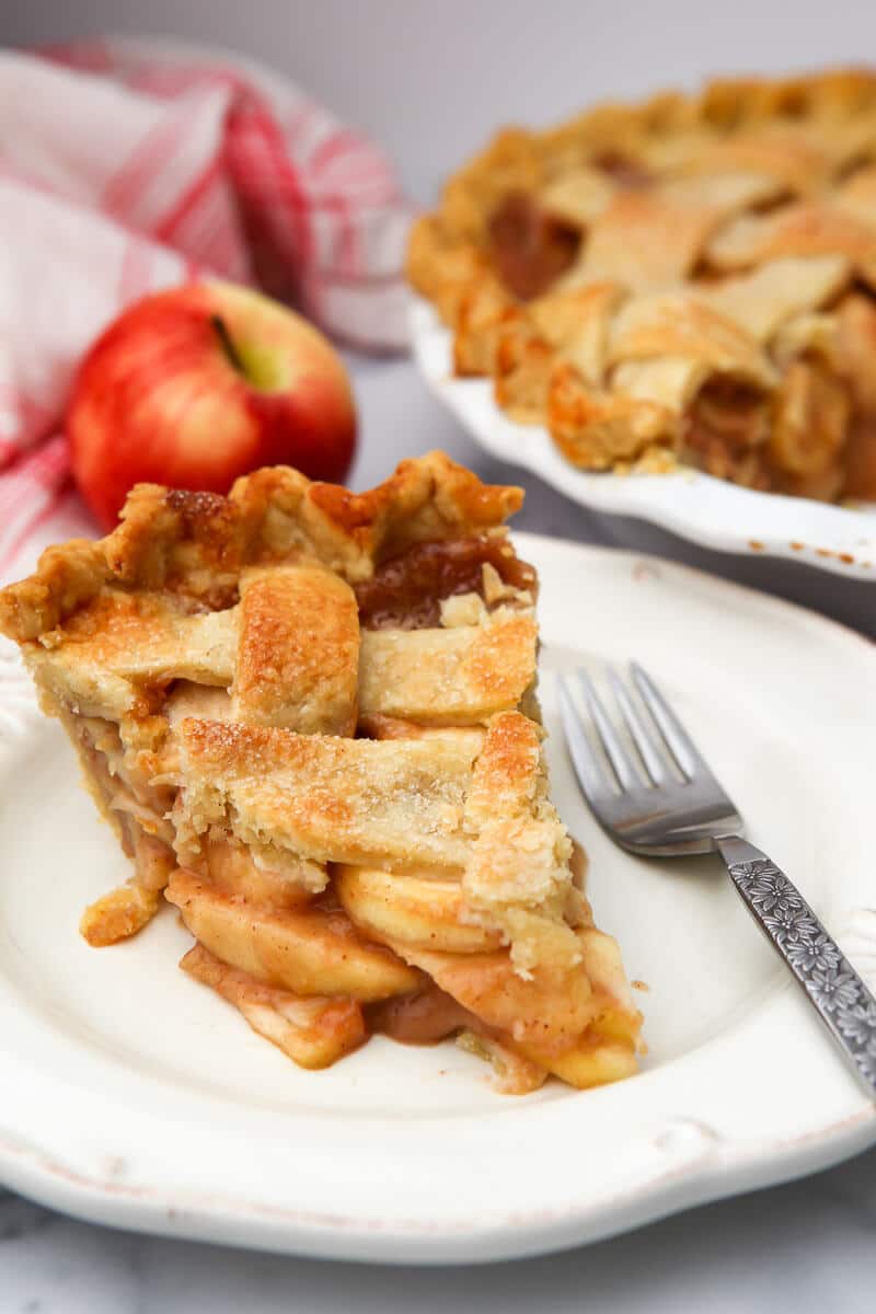 A slice of an apple pie with a full pie and an apple behind it.