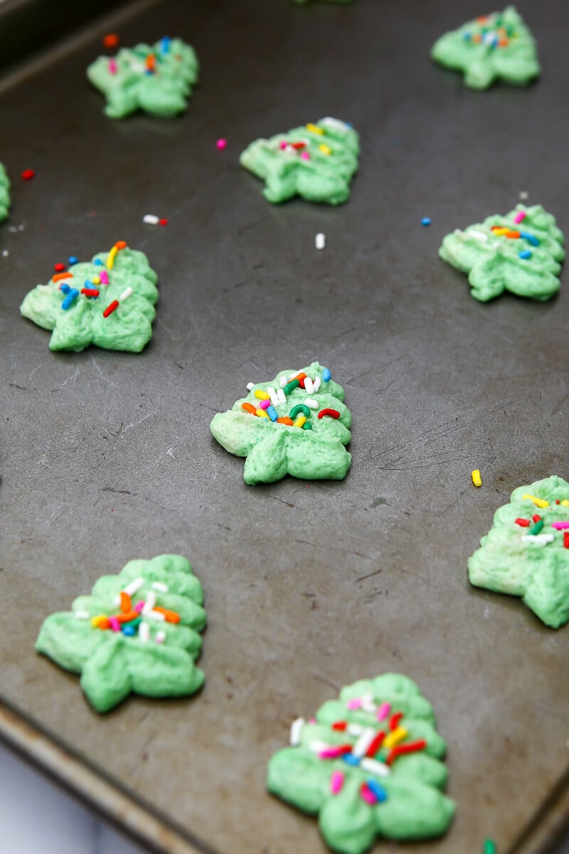 Vegan spritz cookies shaped like Christmas trees and colored green with sprinkles. on a cookie sheet.