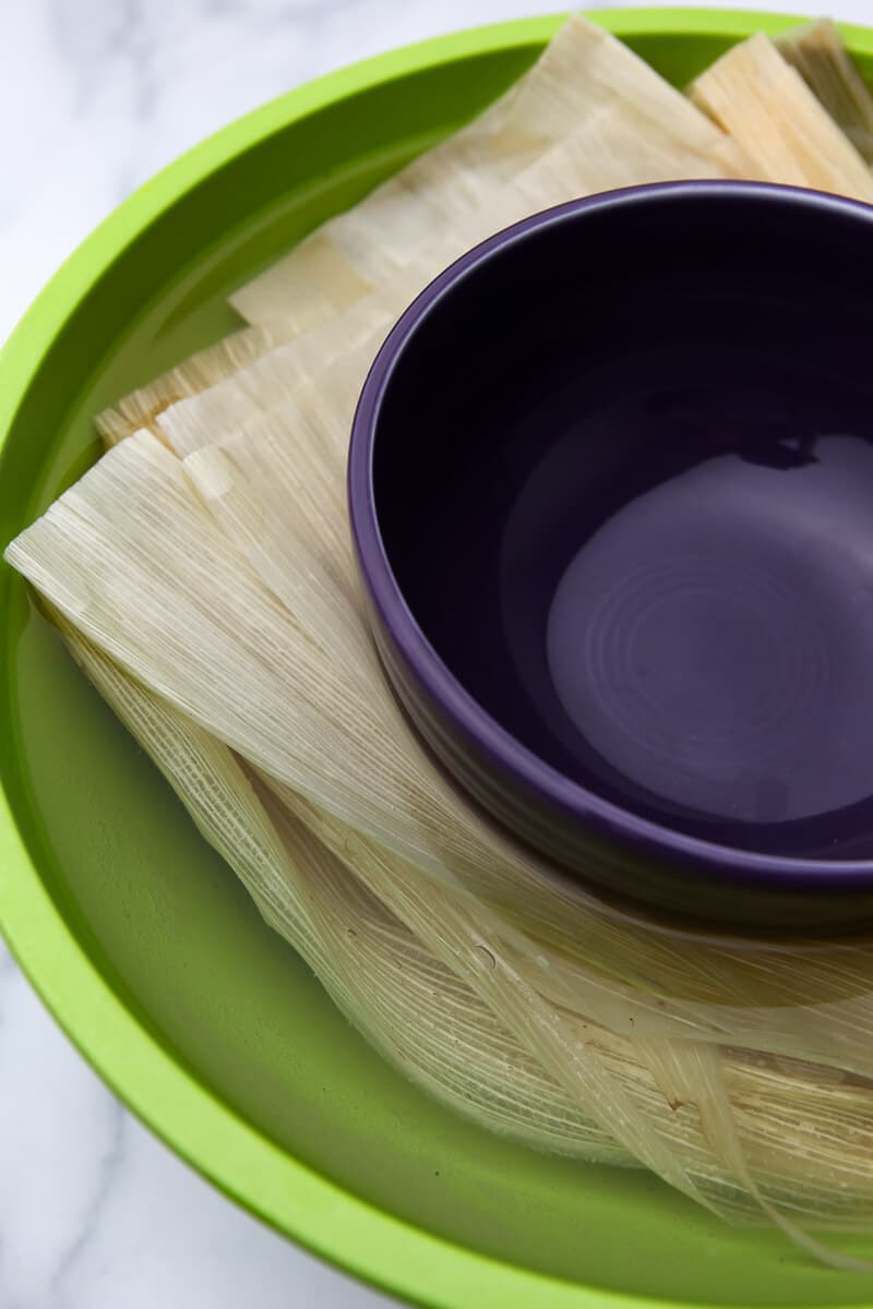 Corn husks soaking in a large bowl of warm water with another bowl on top to weigh them down.