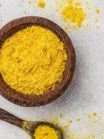 A wooden bowl filled with nutritional yeast with a spoonful on the side.