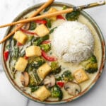 A plate of vegan Thai green curry with tofu with a mound of rice in the middle and chop sticks on the side.