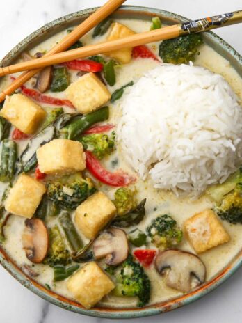 A plate of vegan Thai green curry with tofu with a mound of rice in the middle and chop sticks on the side.