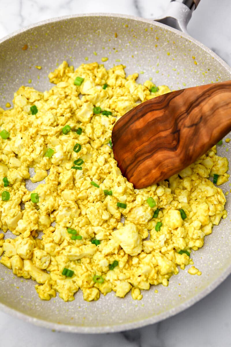 A white frying pan with a vegan tofu scramble made with silken tofu being stirred with a wooden spoon.