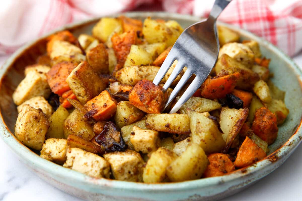 A close up of a plate full of roasted veggies and tofu on a plate with a fork sticking in it.