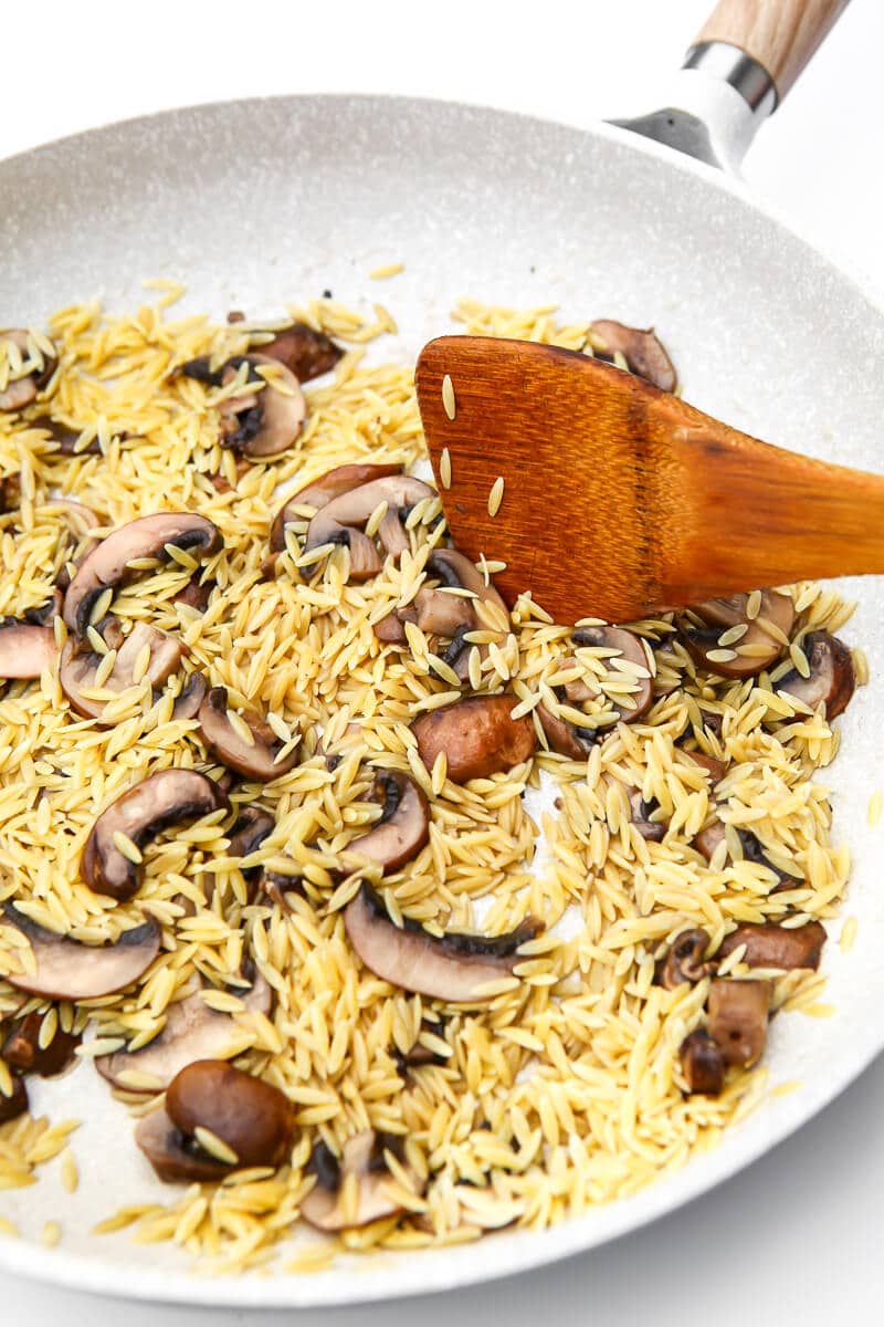 A skillet with sauteed mushrooms and cooked orzo being mixed with a wooden spoon.