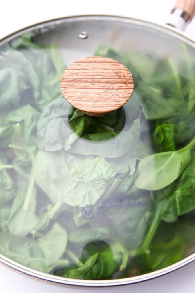 A skillet of vegan orzo with fresh spinach on top, covered with a glass lid.