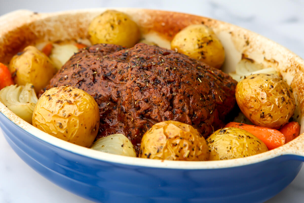 A vegan pot roast in a blue caserole dish with a seitan roast, potatoes, carrots, and onions all drizzled in a basting sauce.