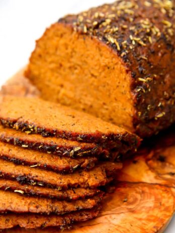 A vegan roast beef seasoned and sliced thin on a wooden cutting board.