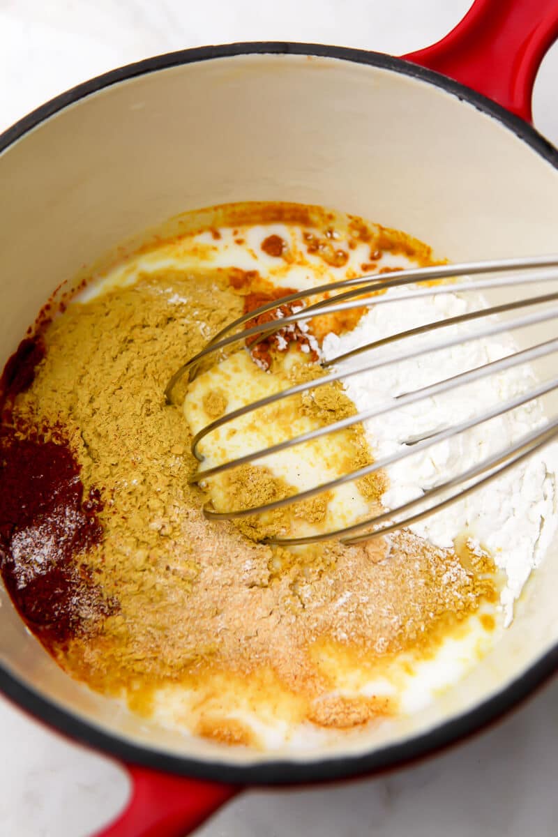 A red saucepan with coconut milk, nutritional yeast, tapioca starch, corn starch, salt and spices to make mac and cheese sauce.