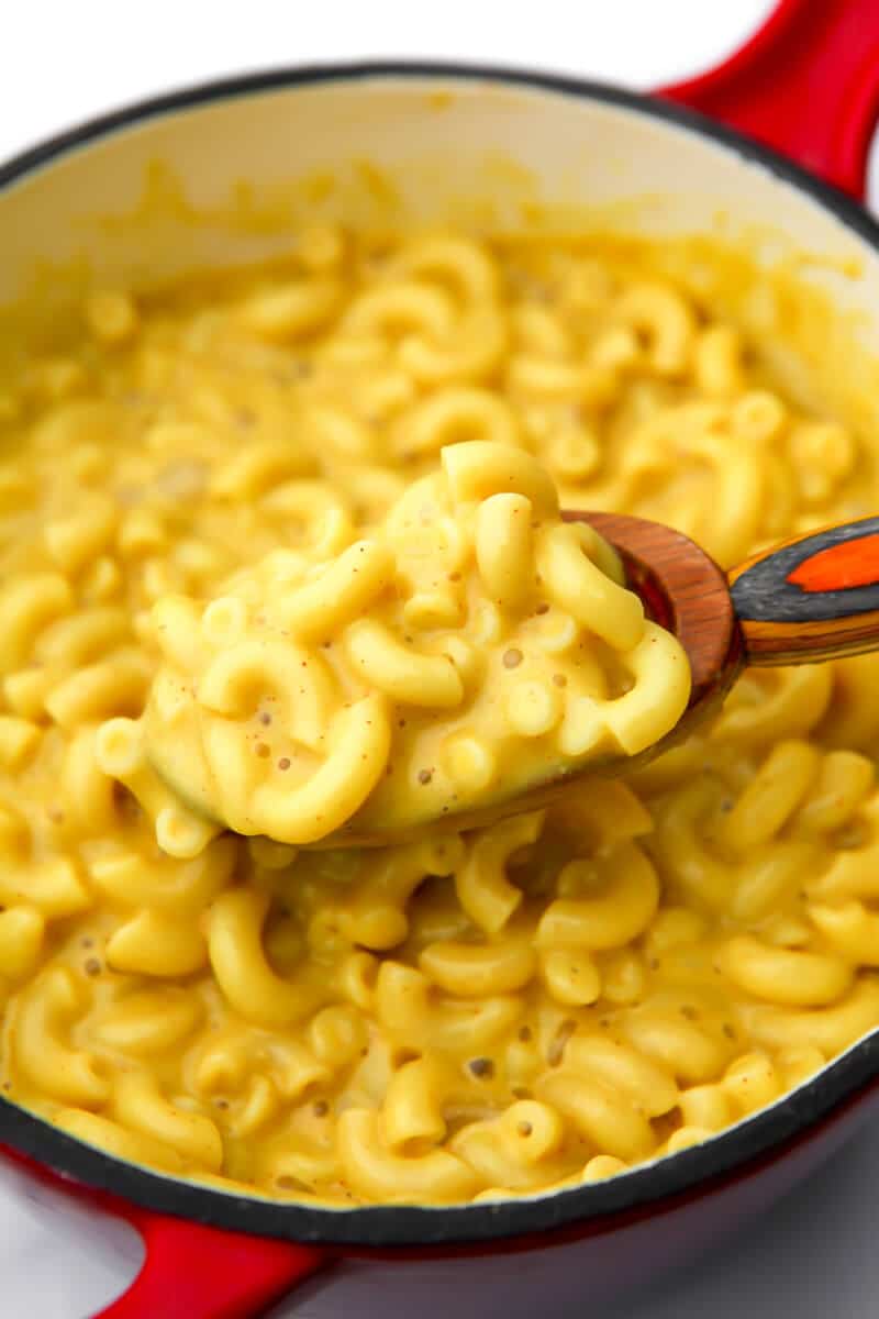 A pot full of creamy coconut mac and cheese with a wooden spoon scooping up a spoonful.