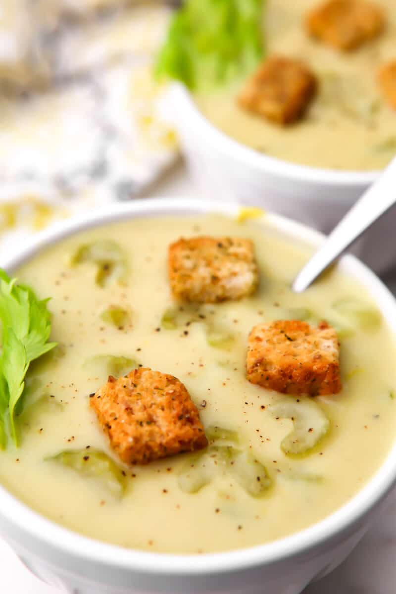 A white bowl filled with vegan cream of celery soup with croutons on top and another bowl of soup behind it.