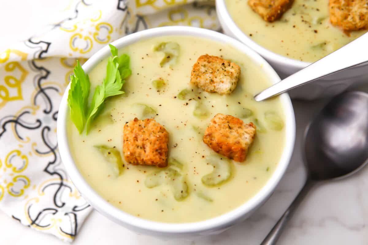 Two bowls filled with creamy vegan celery soup with croutons on the top and a tea towel on the side.