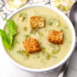 A top view of a a bowl of cream of celery soup with some celery leaves on the side and croutons on top.