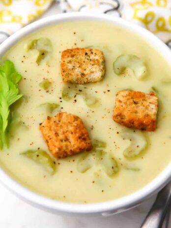 A top view of a a bowl of cream of celery soup with some celery leaves on the side and croutons on top.