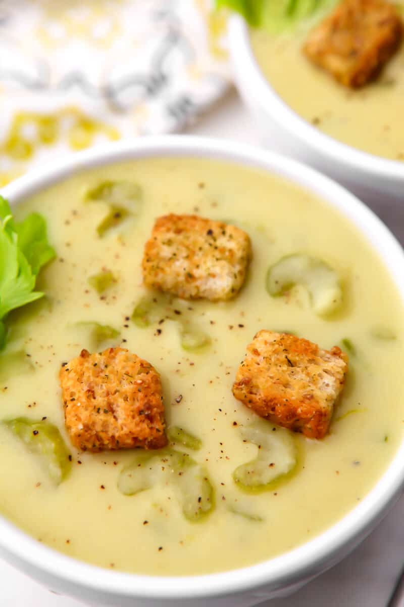 A white bowl filled with vegan cream of celery soup with croutons on top of it.