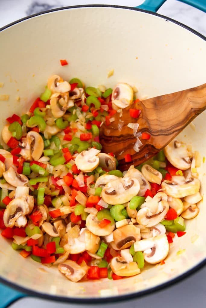 A pot of vegetables sautéing, being stirred by a wooden spoon.