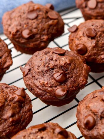 Vegan double chocolate banana muffins on a cooling rack with a blue tea towel behind it.