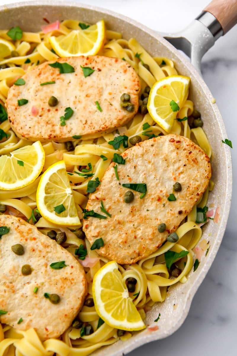A frying pan with vegan piccata with vegan chicken and lemon wedges on top.