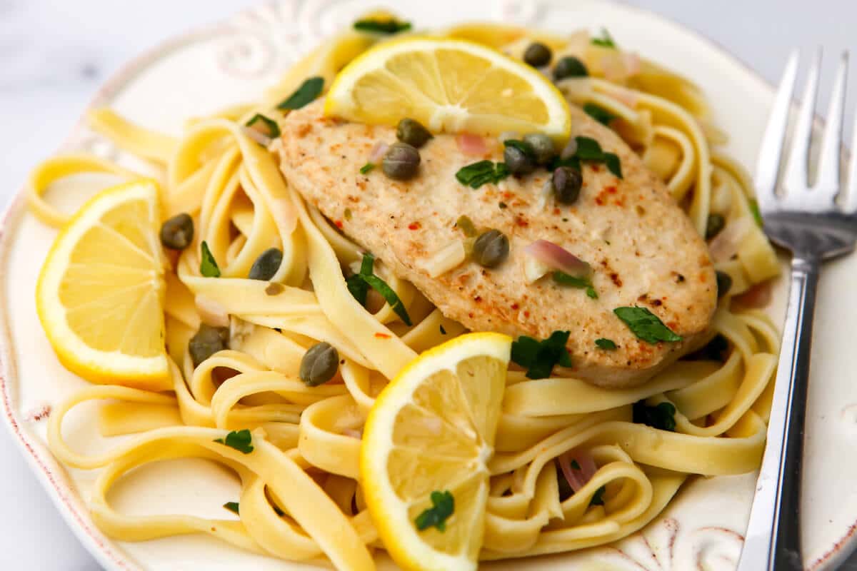 A white plate full of vegan chicken piccata with a fork on the side and lemon wedges around it.