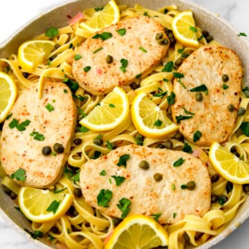 A frying pan filled with vegan piccata with vegan chicken cutlets, capers, and lemon wedges on top.