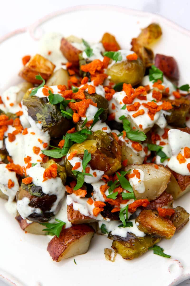 A white plate filled with roasted red potatoes and brussel sprouts topped with vegan ranch dressing and bacon bits.