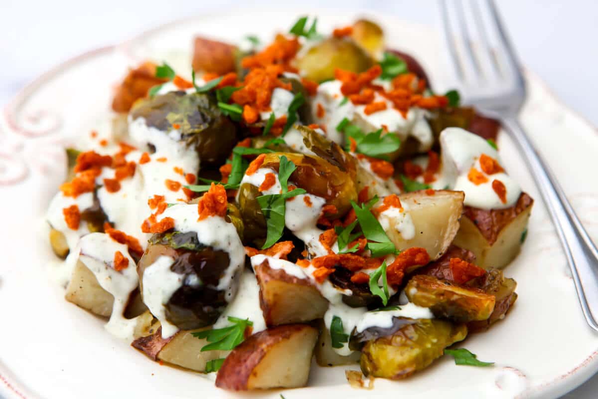 A plate full of roaste potatoes and brussel sprouts topped with vegan bacon bits and vegan ranch dressing.