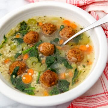A bowl of vegan Italian wedding soup with vegan meatballs with a red and white tea towel next to it.