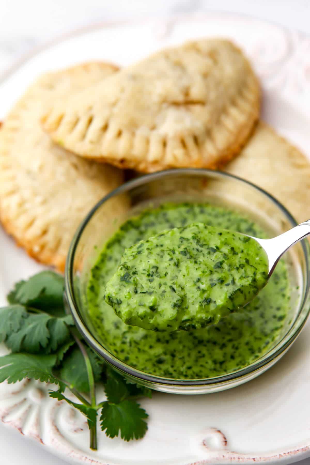 A bowl of garlic cilantro sauce with lime and jalapanos with empanadas behind it. 