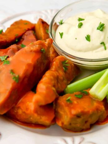 Vegan buffalo chicken wings on a white plate with vegan blue cheese dip and celery on the side.