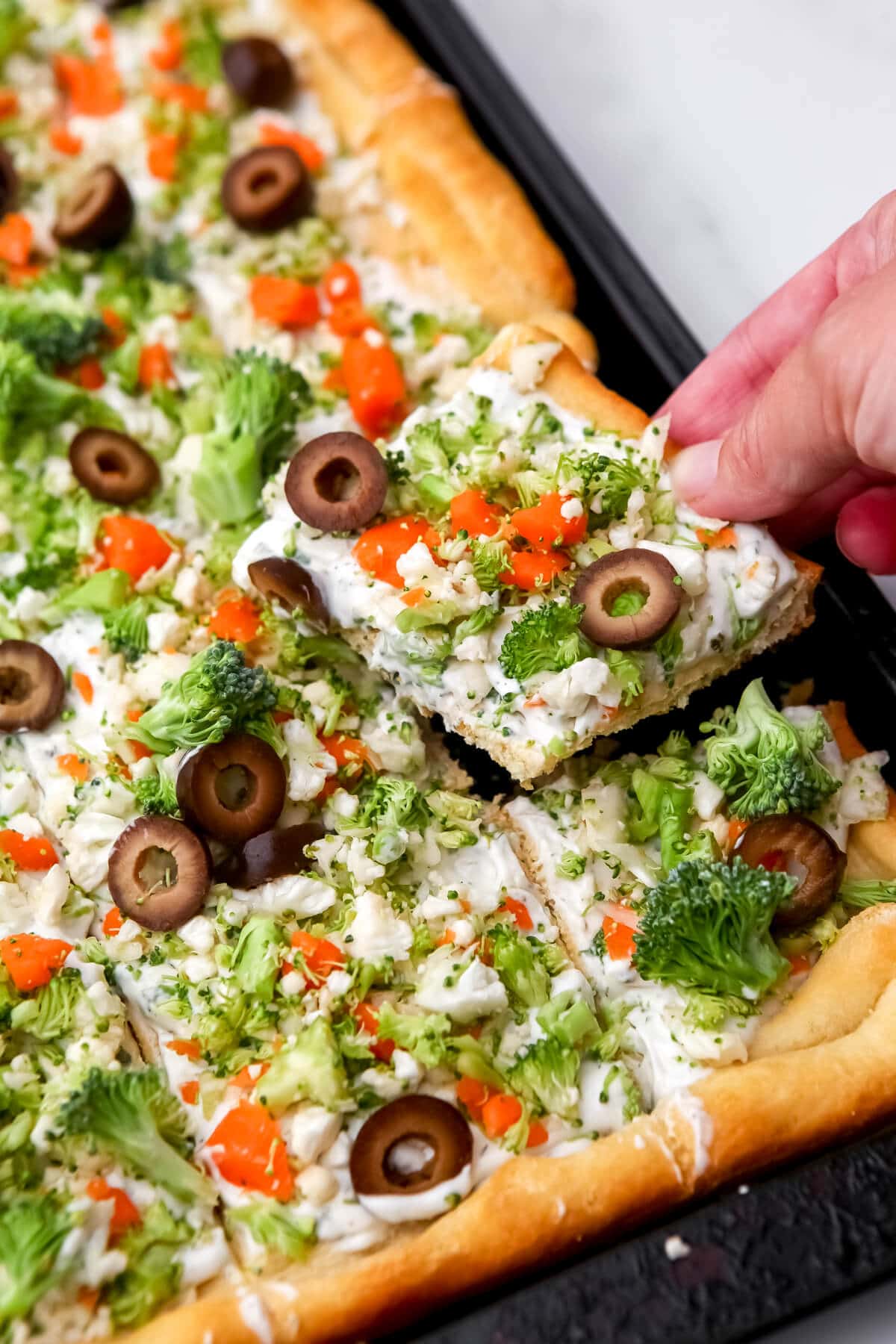 A hand picking up a piece of vegan cold veggies pizza from a tray