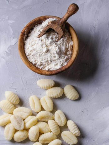 A top view of gnocchi on a blue countertop with a bowl of flour near it.