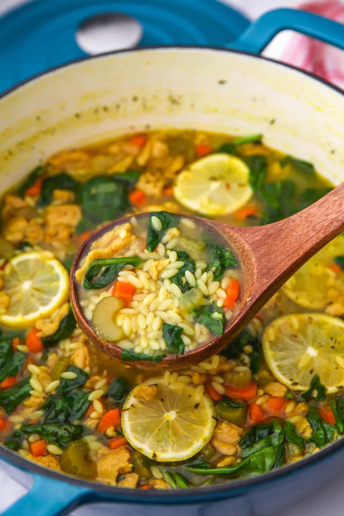A blue dutch oven filled with vegan orzo soup being scooped up with a wooden ladel.