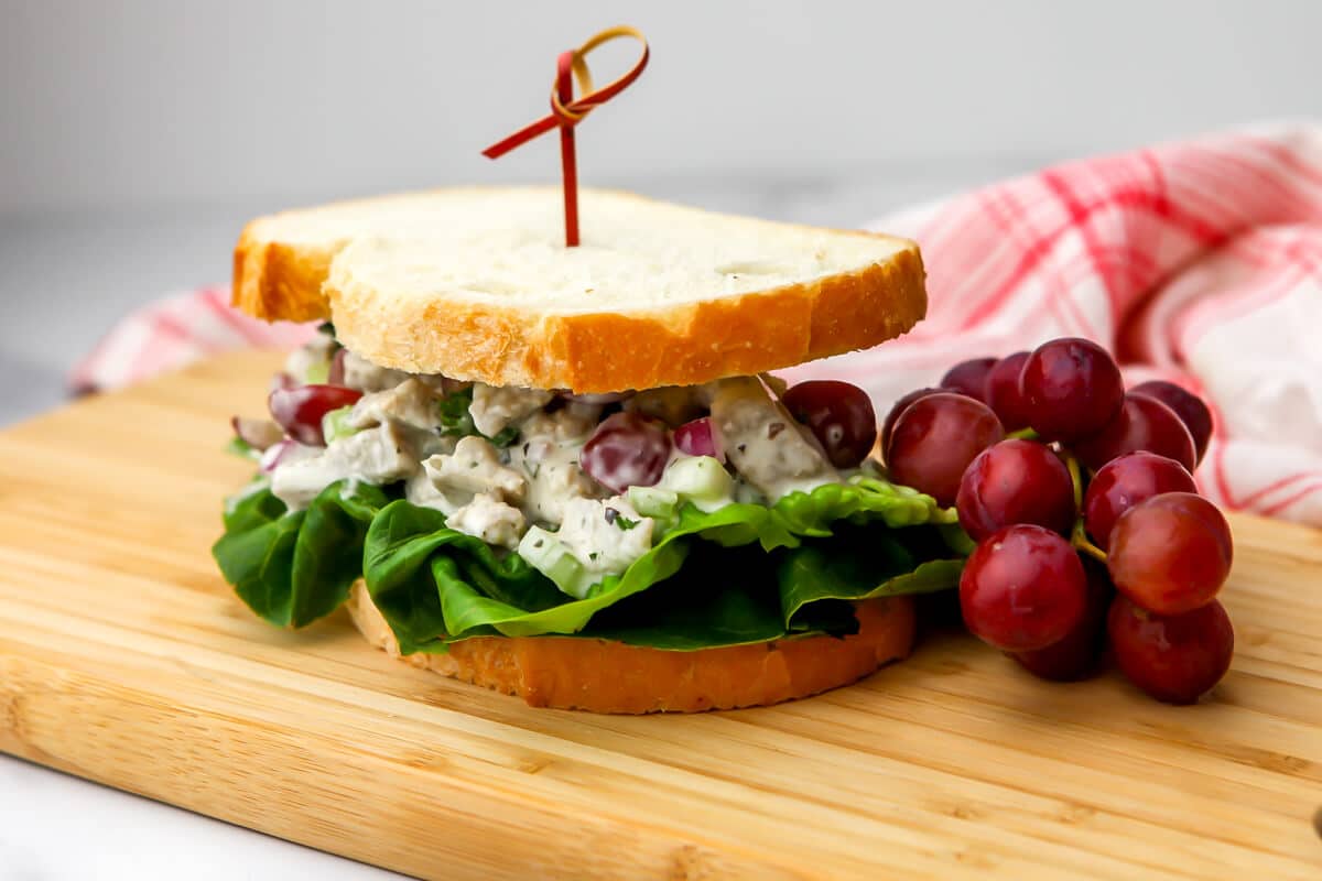 A vegan chicken salad sandwich on a cutting board with grapes on the side.