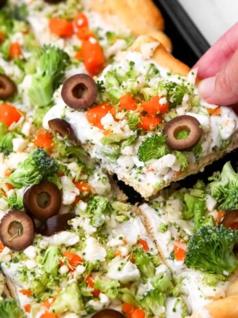 Vegan cold veggie pizza on a cookie sheet with a hand picking up a piece.