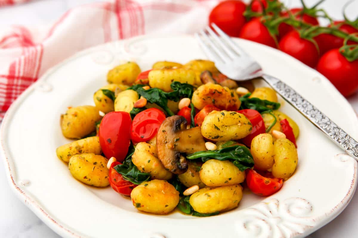 A white plate filled with vegan gnocchi and sauted veggies with tomatoes and a tea towel on the side.