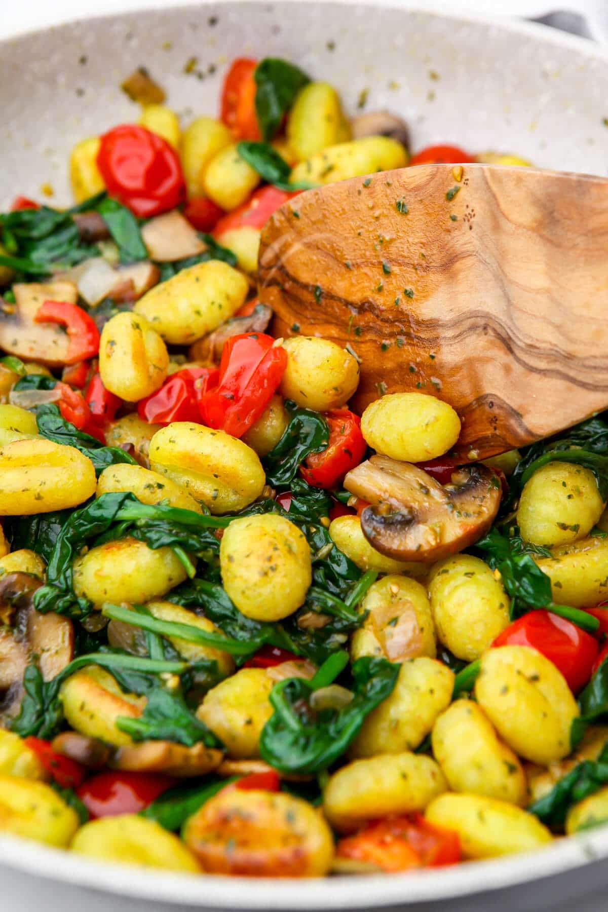 A white frying pan filled with gnocchi, mushrooms, spinach, tomaotes, and pesto.