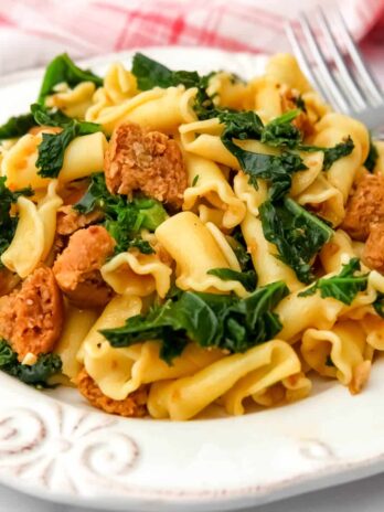 Vegan sausage pasta with kale on a white plate with a fork on the side.
