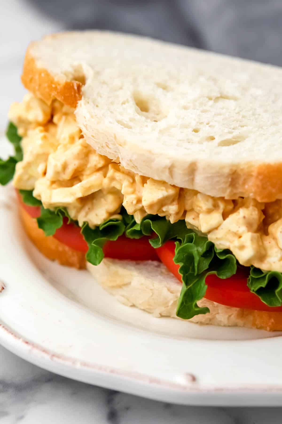 A tofu egg salad sandwich on a white plate with tomatoes and lettuce.