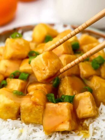 Vegan orange tofu on a plate of white rice with chopsticks picking up a piece.