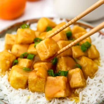 Vegan orange tofu on a plate of white rice with chopsticks picking up a piece.