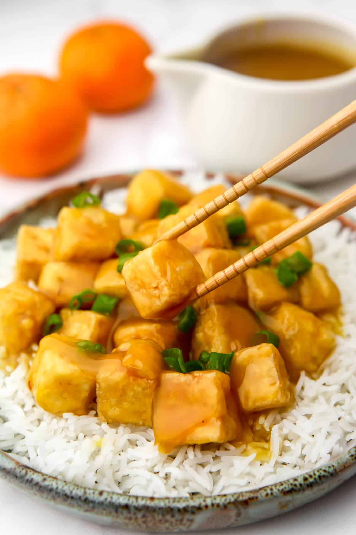 Crispy orange tofu on a bed of white rice with a container of orange sauce and 2 oranges behind it.
