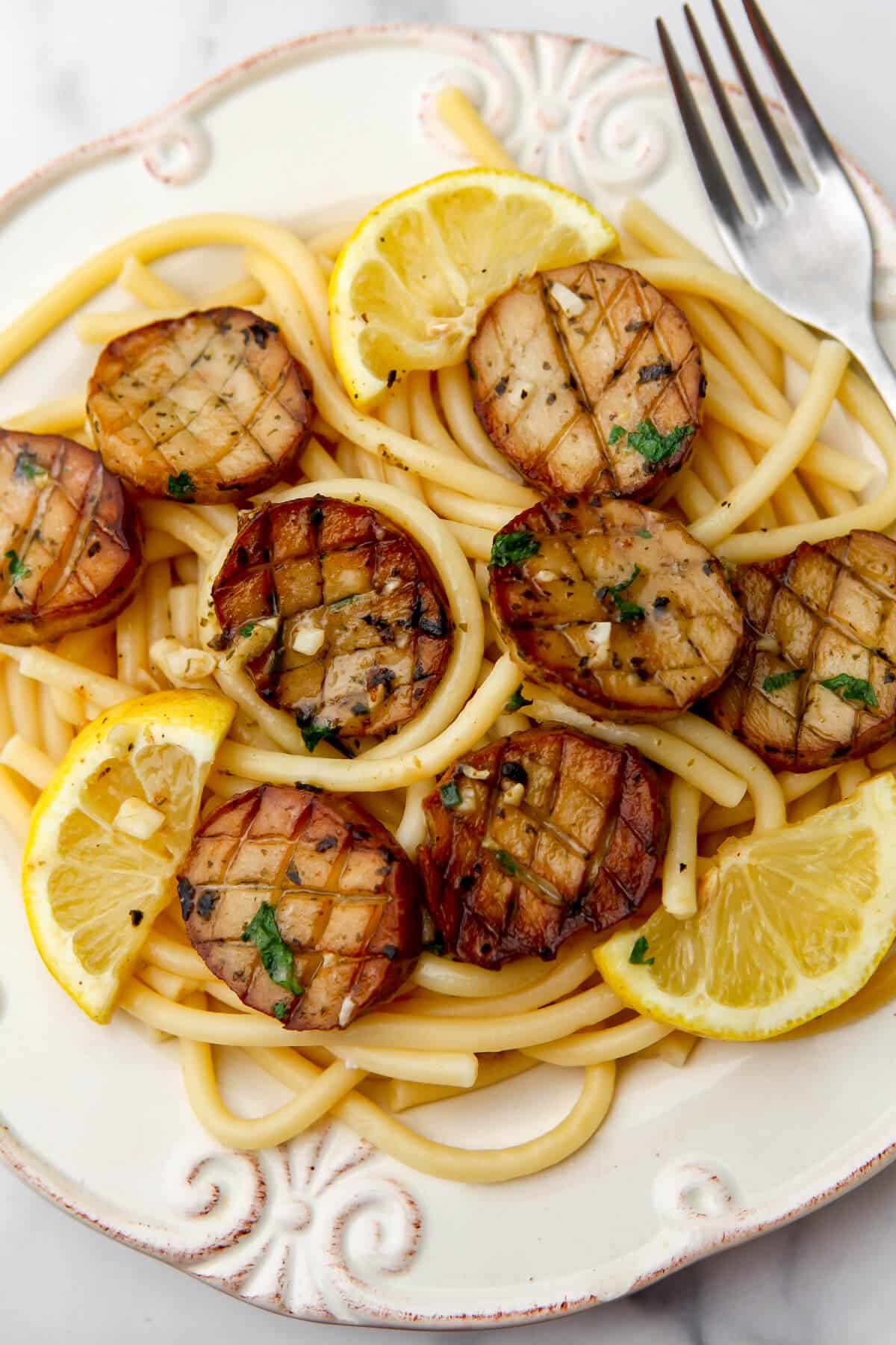 A white plate with vegan scallops in garlic butter sauce served over pasta with wedges of lemon.