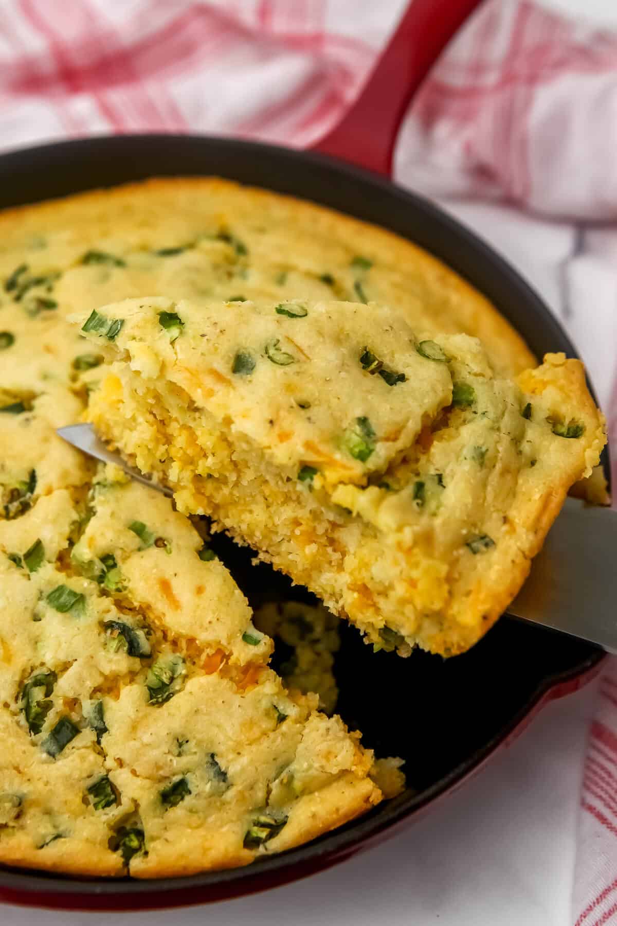 A vegan jalapeno cornbread baked in a red iron skillet with a piece being taken out.