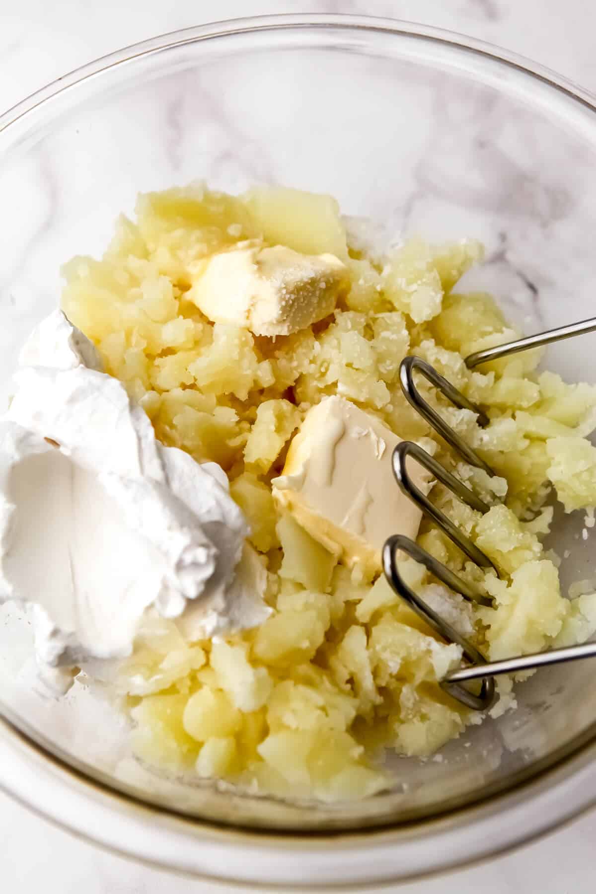 Potatoes, butter, sour cream, and vegan milk in a bowl being mashed.